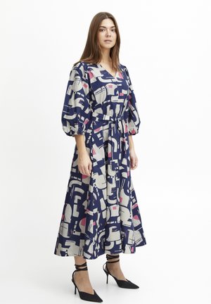 Knielanges Kleid in Marineblau mit abstrakten Mustern in Creme, Pink und Schwarz. Verfügt über einen V-Ausschnitt, taillierten Gürtel und Puffärmel. Trägt schwarze High Heels.