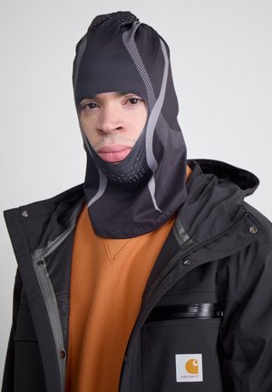 Homme portant une cagoule noire et grise à motifs, un sweat-shirt orange et une veste Carhartt noire avec fermeture éclair et boutons-pression.