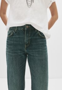 Visoko pasne denim hlače v temno modri barvi z teksturirano površino, vidnim šivom in sprednjimi žepi. V kombinaciji z belo majico s kratkimi rokavi.