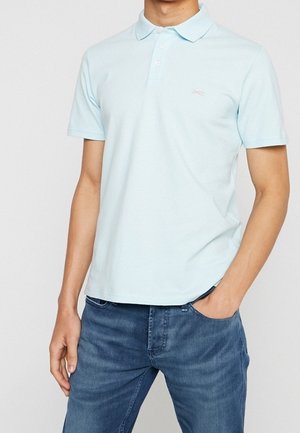 Hombre con camiseta polo de manga corta azul claro con cuello y tres botones, combinada con jeans de mezclilla azul.