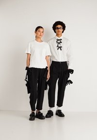 Um homem e uma mulher estão lado a lado, vestindo calças pretas com detalhes em fita, camisas brancas e sapatos pretos, em frente a um fundo branco liso.