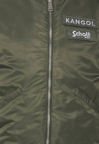 Casaco bomber verde-oliva com fecho prateado, dois bolsos laterais e remendos de tecido rotulados "Kangol" e "Schott NYC." Textura suave.