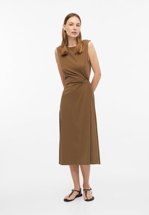Frau trägt ein ärmelloses, braunes Midikleid mit gerafftem Detail an der Taille und schwarzen flachen Sandalen, steht vor weißem Hintergrund.