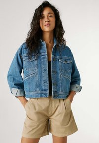 Ung kvinde med langt krøllet hår iført en blå denimjakke over en sort top og beige shorts, stående med hænderne i lommerne.