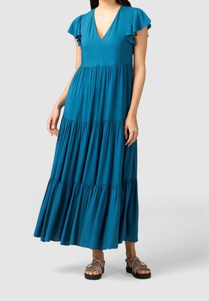 Vrouw die een teal maxi-jurk met tierlagen en vleugelmouwen draagt, met zwarte plateauzandalen met gevlochten banden, staande op een lichtgrijze achtergrond.