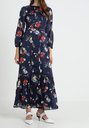 Donna che indossa un lungo abito floreale blu navy con fiori rossi, bianchi e gialli e tacchi bianchi a punta con cinturini alla caviglia.