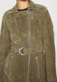 Veste en daim vert olive avec une taille ceinturée, des poches zippées et un col à boutons-pression. Elle présente une texture lisse et un design ajusté.
