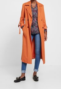 Oranger Trenchcoat mit tailliertem Schnitt, doppelreihigem Design und großen Revers. Kombiniert mit einer gemusterten Bluse, blauen Jeans und schwarzen Mokassins.