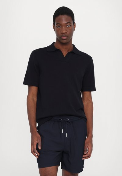 Polo preto de manga curta com colarinho, apresentando um design em forma de V. Usado com calções curtos de cordão azul-marinho. Textura de tecido suave.