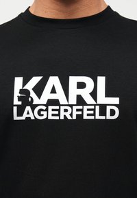 Μαύρο μπλουζάκι με μεγάλο λευκό κείμενο "KARL LAGERFELD" και μια σιλουέτα άνδρα με γυαλιά ηλίου και αλογοουρά μέσα στο γράμμα "K".