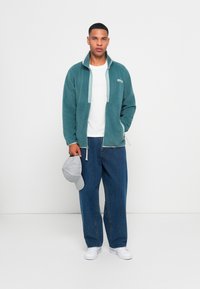 Teal fleecejakke med halv-zip design, lyseblå detaljer, båret over en hvit t-skjorte og kombinert med løse blå jeans og en grå caps.