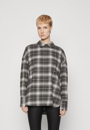 Camicia a quadri oversized nera e bianca con chiusura a bottoni, colletto a punta e maniche lunghe, caratterizzata da un orlo leggermente curvo.