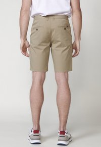 Shorts khaki dintr-un amestec de bumbac. Prezintă două buzunare pe spate cu nasturi. Purtați cu un tricou alb și adidași gri cu accente roșii.