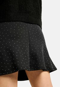 Rozšířená černá sukně s hladkou strukturou, zdobená drobnými bílými puntíky po celé ploše a jemně se rozšiřující lemem.