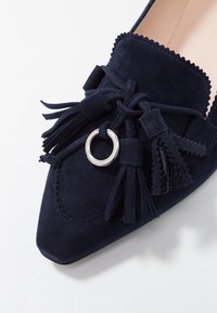 Marineblauwe suède platte schoen met een spitse neus, decoratieve kwastjes, een zilveren ring-accent en gegolfde rand rondom de kraag.