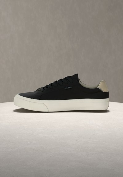 Baskets basses noires avec semelle blanche et languette de talon beige, présentées sur une surface claire sur fond neutre.