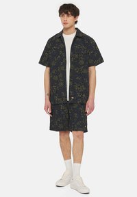 Completo di camicia a maniche corte e pantaloni shorts di colore blu scuro con un motivo astratto verde, realizzato in tessuto leggero; abbinato a sneaker bianche e calzini.