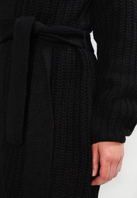 Knitted black cardigan with a textured pattern, featuring a tied belt at the waist and long, voluminous sleeves.