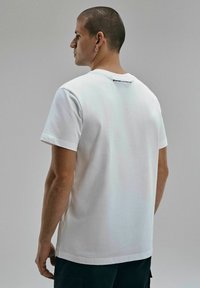 Biała bawełniana koszulka z krótkim rękawem, okrągłym dekoltem i logo z tyłu. Gładka faktura, luźny krój i średnia długość.