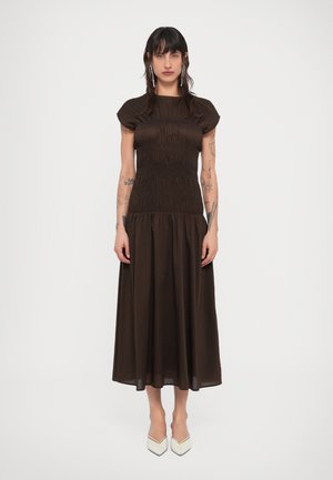 Woman with dark hair wearing long, dark brown ruched dress, white pointed shoes, and dangling earrings against plain white background.