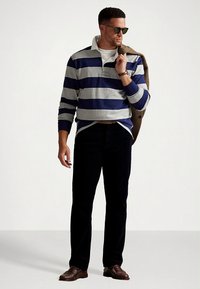 Polo de manga longa às riscas azul-marinho e cinzento com gola branca, combinado com calças escuras e sapatos castanhos, complementado com óculos de sol e um casaco.