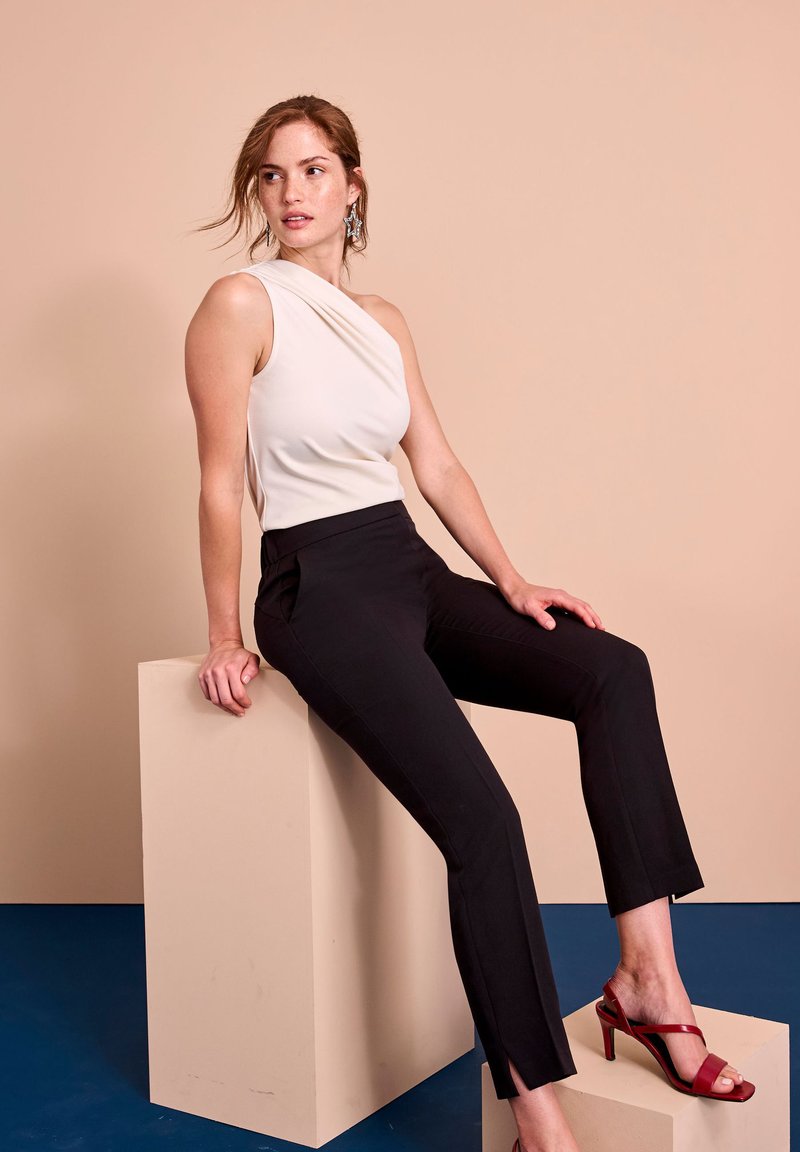 Witte top met één schouder, zwarte op maat gemaakte broek met zijsplitten, en rode open teenhakken. Model zit op een beige blok tegen een lichte achtergrond.
