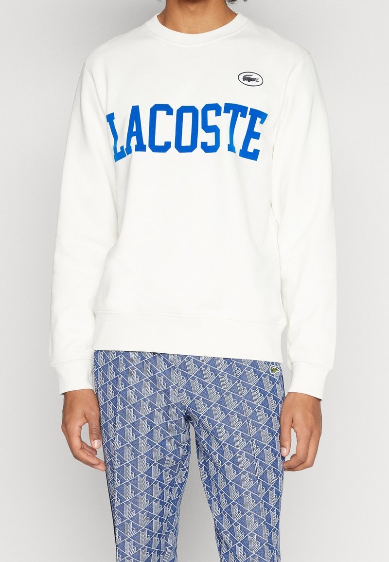 Vit sweatshirt med blå "LACOSTE" bokstäver, med rund hals och ribbad ärm. Matchad med mönstrade blå byxor.