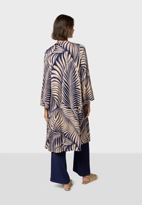 Lång kimono i beige med mörkblå bladmönster, med en lös passform, vida ärmar och rak fåll. Matchas med mörkblå vida byxor.