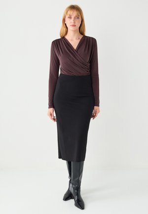 Donkere, bruinachtige wikkeltrui met lange mouwen gecombineerd met een zwarte, nauwsluitende midi-rok en kniehoge zwarte laarzen met hak. Gladde stof met minimale textuur.