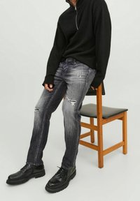 Svart zip-up tröja, grå rivna denimjeans och svarta snör stövlar. Visuell textur i tyget och kontrasterande färgtoner. Trä stol närvarande.