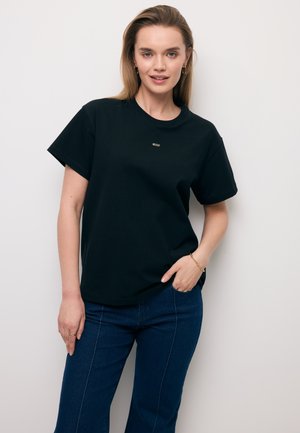 Vrouw met blond haar draagt een zwart T-shirt, donkerblauwe jeans, gouden creooloorbellen, armband en ring, poserend tegen een effen lichte achtergrond.