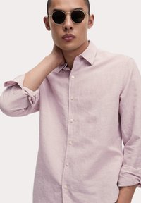 Camisa de color rosa claro, con botones y un patrón texturizado, mangas arremangadas y cuello amplio. Presenta pequeños botones crema y una tela suave.