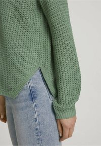 Ljusgrön stickad tröja med ett texturerat mönster, sidoinsnitt och ribbade ärmslut, tillsammans med ljusblå jeans.