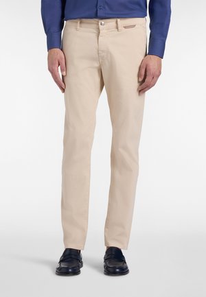 Mężczyzna w beżowych dopasowanych spodniach, granatowej koszuli wizytowej i czarnych eleganckich butach, stojący z ramionami swobodnie opuszczonymi wzdłuż ciała.