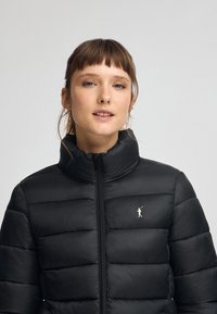 Czarna kurtka puchowa z wysokim kołnierzem, pikowaną konstrukcją i błyszczącym wykończeniem. Posiada subtelne logo na lewej piersi oraz pełny zamek z przodu.