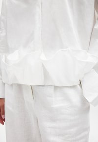White textured blouse featuring a ruffled hem and long sleeves, paired with white linen pants. Smooth fabric with a lightweight appearance.