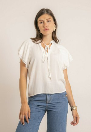 Jeune femme aux cheveux longs bruns portant un chemisier blanc à manches courtes avec un col à nouer et un jean bleu, posant devant un fond beige uni.