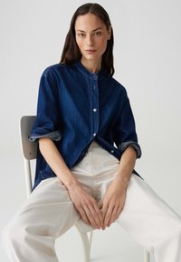 Camicia in denim con chiusura a bottoni, colletto rotondo e maniche arrotolate; di colore blu scuro, abbinata a pantaloni larghi di colore chiaro.