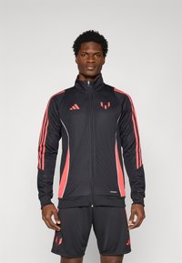 Chaqueta deportiva negra con rayas y acentos rojos, con cierre completo, cuello alto y logo. Combinada con pantalones cortos negros.