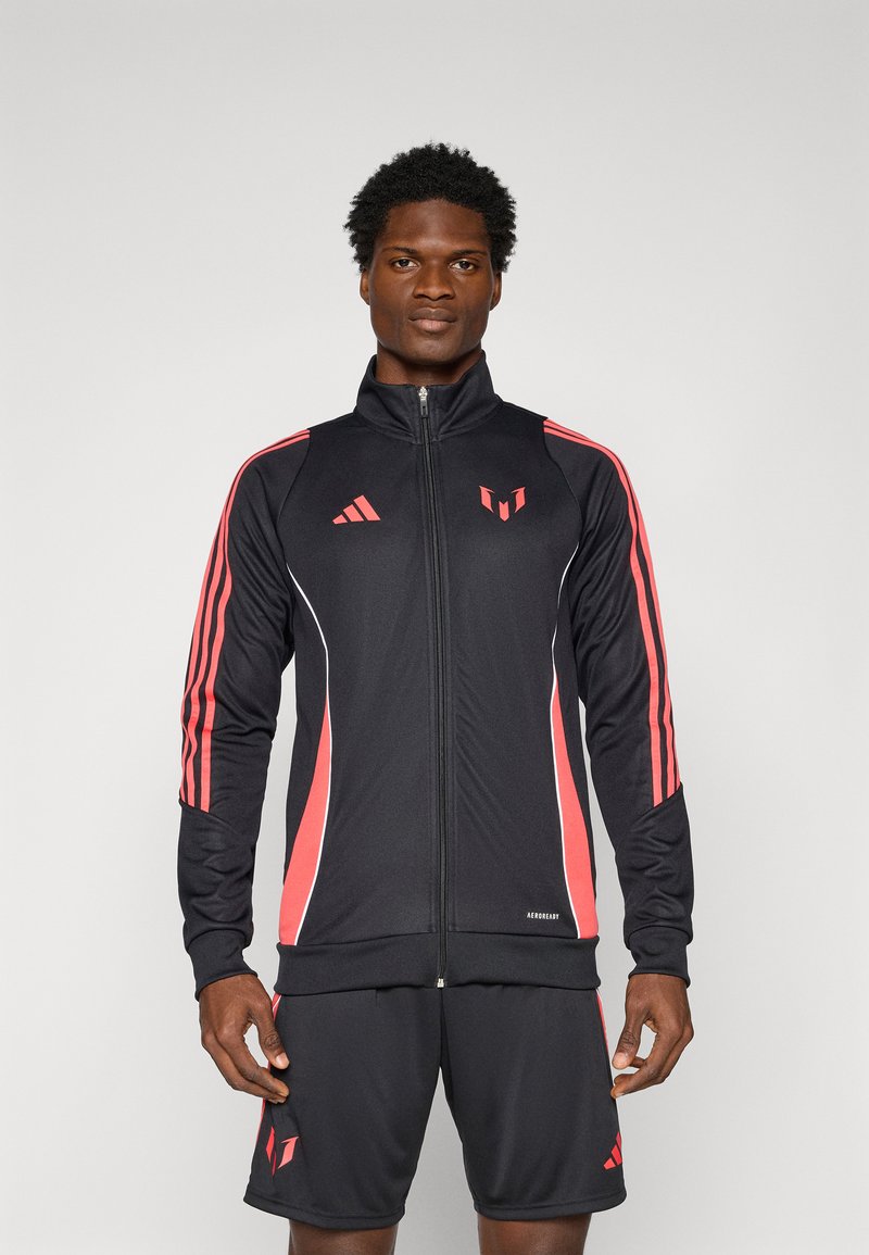Chaqueta deportiva negra con rayas y acentos rojos, con cierre completo, cuello alto y logo. Combinada con pantalones cortos negros.