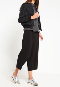 Veste bomber noire avec deux poches avant, haut gris, pantalon noir à large jambe et chaussures argentées brillantes, créant un look élégant et coordonné.