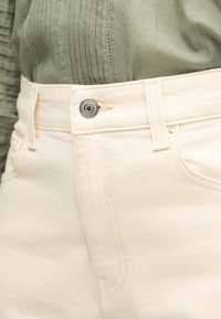 Κοντινό πλάνο στη μέση τζιν παντελονιού από εκρού τζιν ύφασμα με ασημένιο κουμπί και πράσινο ριμπ τοπ μέσα από το παντελόνι.