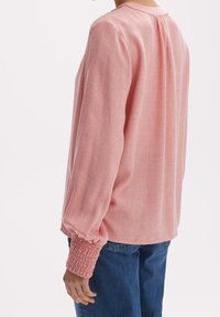 Blouse rose à manches longues en tissu texturé, avec poignets froncés, encolure ronde et coupe décontractée avec un pli au dos.
