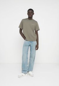 Πράσινο-ελαιί χρώμα βαμβακερό t-shirt με κοντά μανίκια, συνδυασμένο με ανοιχτό μπλε τζιν παντελόνι με ίσια γραμμή και λευκά αθλητικά παπούτσια με γκρι λεπτομέρειες.