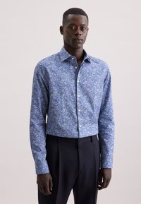 Uomo che indossa una camicia a maniche lunghe blu a fiori infilata nei pantaloni neri, in piedi contro uno sfondo chiaro e neutro.