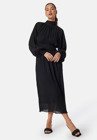 Robe noire à manches longues avec un col haut, un design plissé et une taille élastique. Le tissu transparent présente une silhouette fluide et un ourlet en forme de vagues.