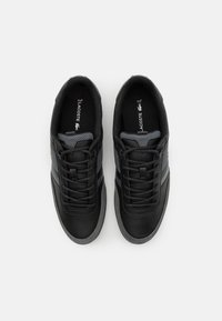 Pár černých tenisek Lacoste s šedými síťovinovými akcenty, černými šňůrkami a šedými podrážkami, zobrazených shora na bílém pozadí.