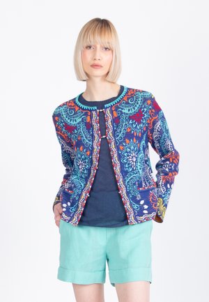 Blond naine kannab mustrilist värvilist jopet üle tumesinise tipu ja hele-türkiissiniste lühikeste pükstega, seisab taskutes käed valgel taustal.