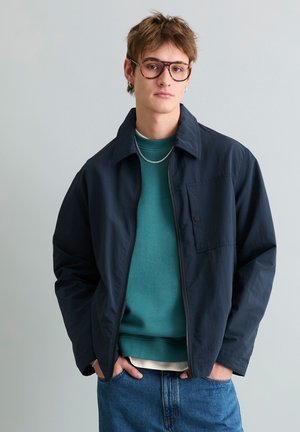 Ung mand med kort brunt hår iført store briller, en mørk marineblå jakke, teal sweatshirt, blå jeans og sølvkæde.