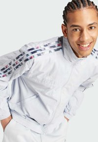 Hellblaue Zip-Up-Jacke mit Streifen, die ein glänzendes geometrisches Muster an den Ärmeln, elastische Bündchen und Fronttaschen aufweist. "adidas"-Logo sichtbar.